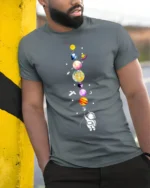 Cute Astronaut Holding Planets Balloon Galaxy T-Shirt - model in grey shirt leaning against column