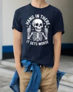 Hang In There It Gets Worse Funny Skeleton Dark Humor T Shirt - model wearing navy tee with hands in pockets