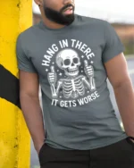 Hang In There It Gets Worse Funny Skeleton Dark Humor T Shirt - model wearing grey tee, leaning on column
