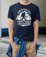 Undefeated Hide And Seek Champion Bigfoot Funny Sasquatch T Shirt - model wearing navy tee with hands in pockets
