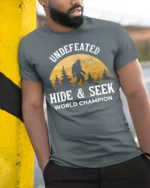 Undefeated Hide And Seek World Champion Bigfoot Funny Sasquatch T Shirt - model wearing grey tee, leaning on column
