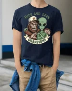 Hide And Seek Champions Funny Alien Bigfoot Graphic T Shirt Style - model wearing navy tee with hands in pockets