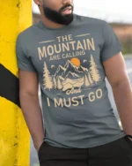 The Mountains Are Calling And I Must Go Hiking T Shirt Adventure Tee - model wearing grey tee, leaning on column