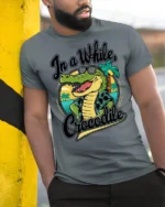 In A While Crocodile Funny Vacation Graphic Tee Beach Humor - model wearing grey tee, leaning on column