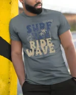 Surf Vibe Ride Wave Graphic Tee Retro Beach Adventure Style - model wearing grey tee, leaning on column