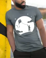 Boy Flying Kite Moonlight Silhouette Graphic T Shirt Dreamy Tee - model wearing grey tee, leaning on column