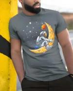 Creative Astronaut Riding Crescent Moon Space Graphic Tee - model wearing grey tee, leaning on column