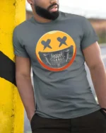 Striking Yellow Smiley Face Skull Mask Graphic Tee - model wearing grey tee, leaning on column